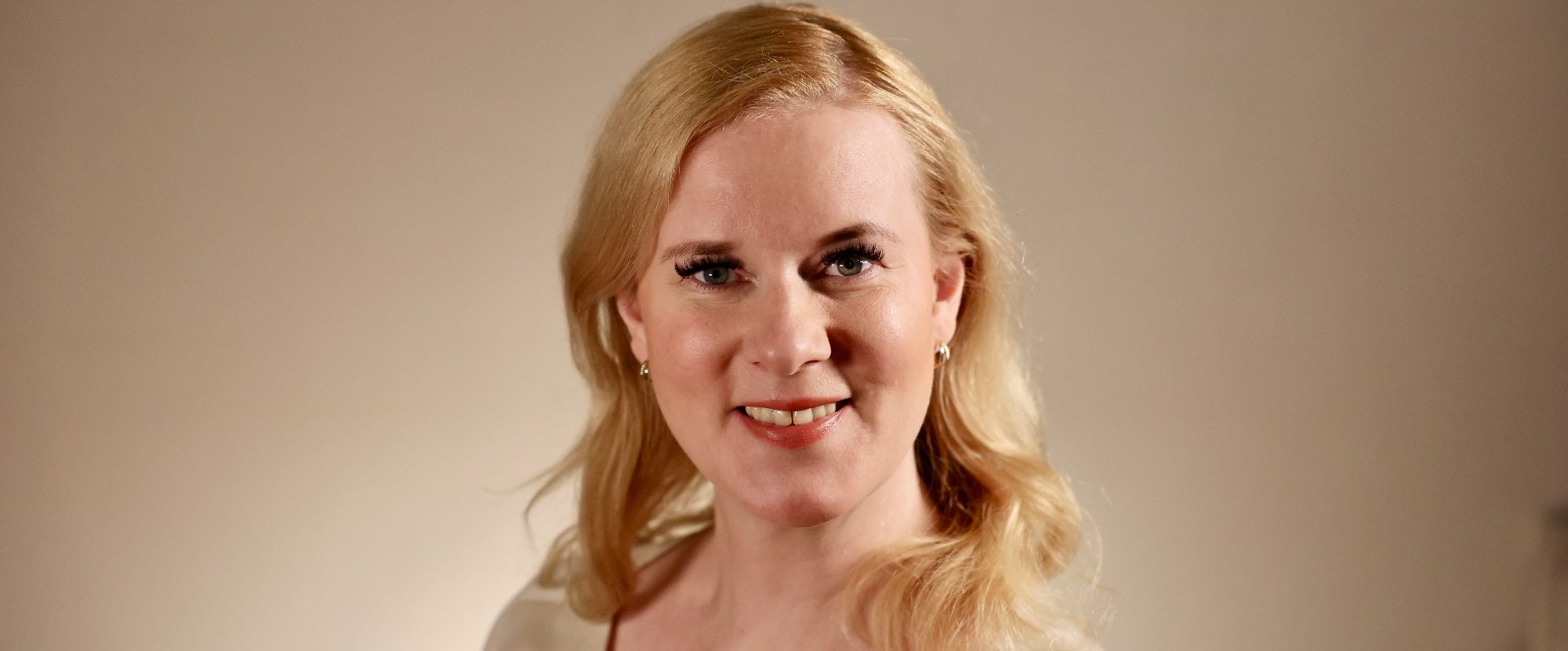 Lächelnde Frau mit blonden Haaren und hellem Oberteil vor neutralem Hintergrund.