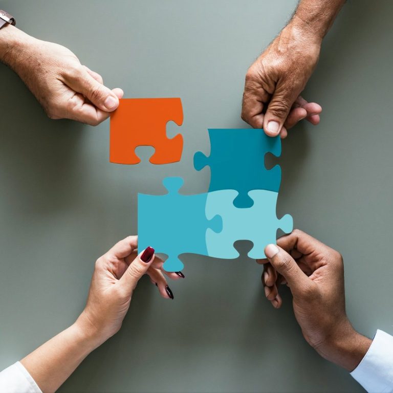 Hände halten orange und türkisfarbene Puzzlestücke über einem grauen Tisch.